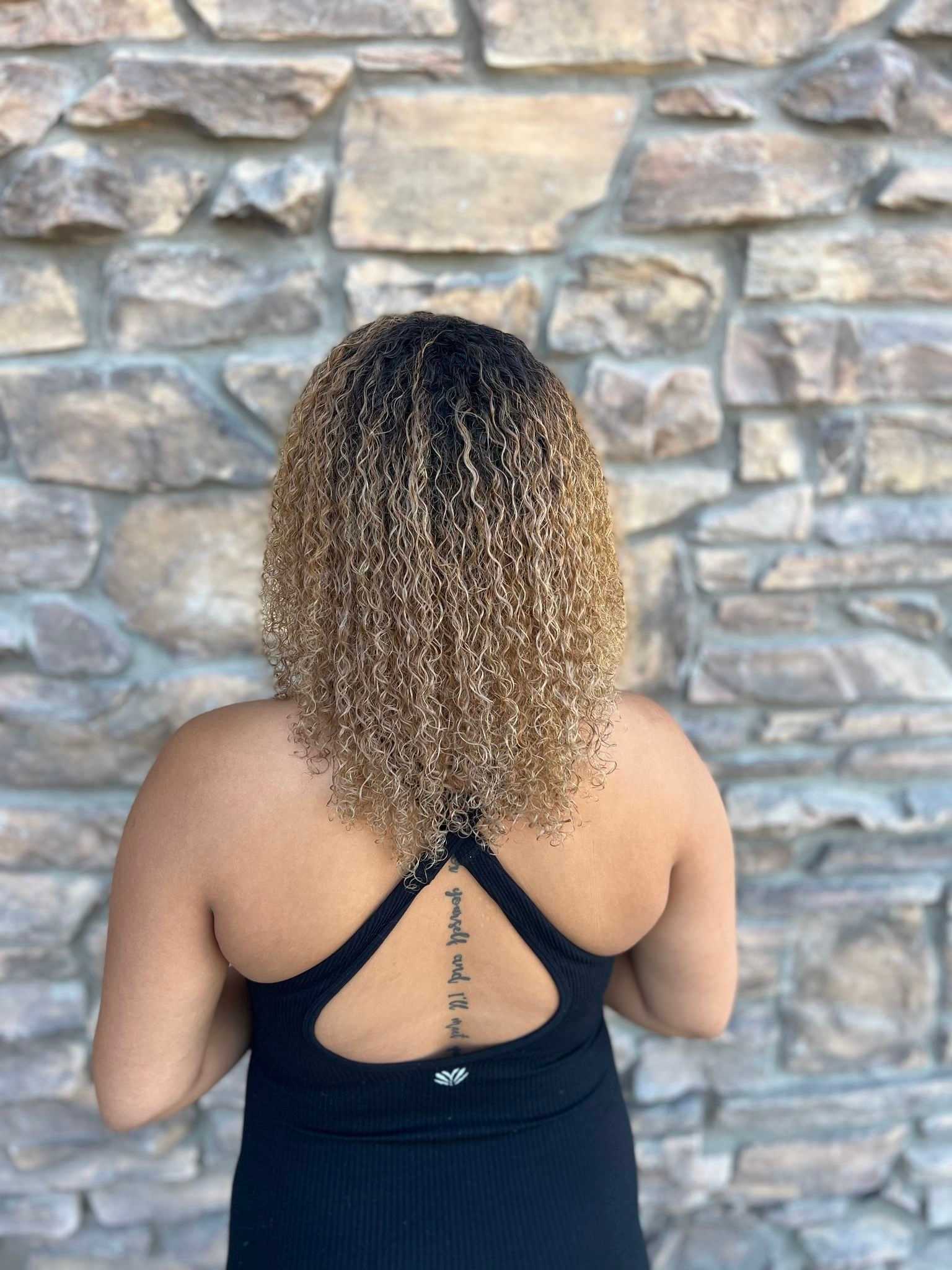 Back view of a person with curly hair and a tattoo, standing against a stone wall.