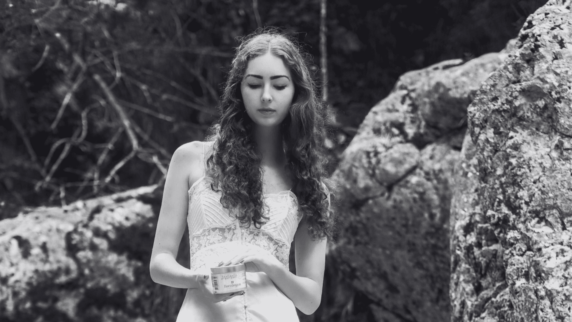 Woman in white dress holding a jar, standing outdoors against rocky background in black and white.